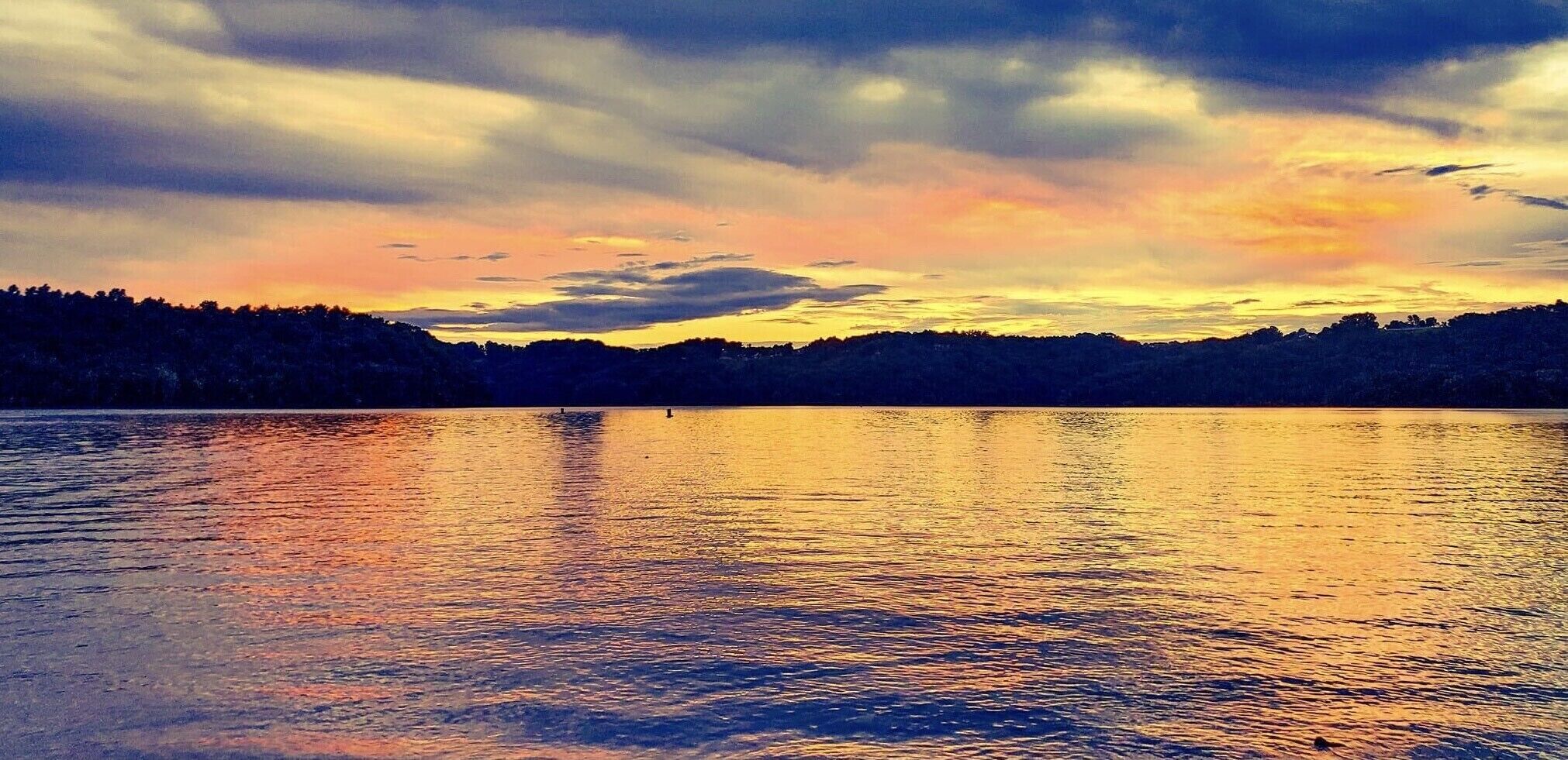 Un lac paisible au coucher du soleil, avec en arrière-plan la silhouette d’une colline boisée.