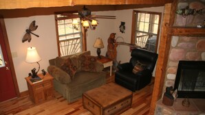 A cozy living room with a wooden floor, a leather armchair, a sofa, a small table with a lamp, and a fireplace.