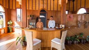 A wooden reception desk with two wicker chairs, a ceiling fan, and potted plants. - Inle Heritage Stilt House (Nyaungshwe)