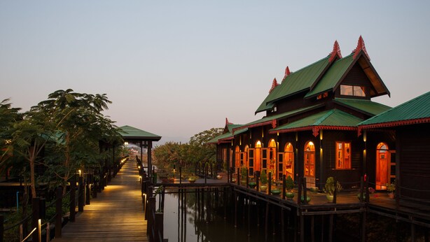 A wooden walkway leading to a traditional building with a green roof, surrounded by water and palm trees. - Inle Heritage Stilt House (Nyaungshwe)