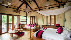 A spacious room with a large bed, a smaller bed, and a daybed. There is a balcony with a wooden deck and a view of greenery. - Inle Heritage Stilt House (Nyaungshwe)