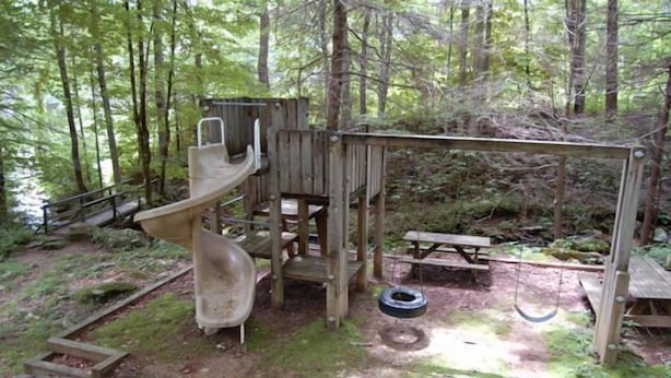 Une structure de jeux en bois, comprenant un toboggan et une balançoire, située dans une zone boisée.
