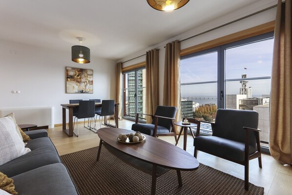 A modern living room with a sofa, a dining table with chairs, and a large window offering a city view. - Expo Panoramic View by Homing (Lisbon)
