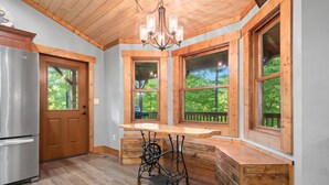A kitchen with wooden cabinetry, a stainless steel refrigerator, a wooden bench, and large windows with a view of trees. - 2019 Newly Built ~ LUXURY Cabin ~ 23 Acre Estate ~ Hot Tub ~ 8 miles to Downtown (Asheville)