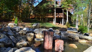 A rustic cabin with a wooden deck, surrounded by a natural landscape with a pond and large rocks. - 2019 Newly Built ~ LUXURY Cabin ~ 23 Acre Estate ~ Hot Tub ~ 8 miles to Downtown (Asheville)