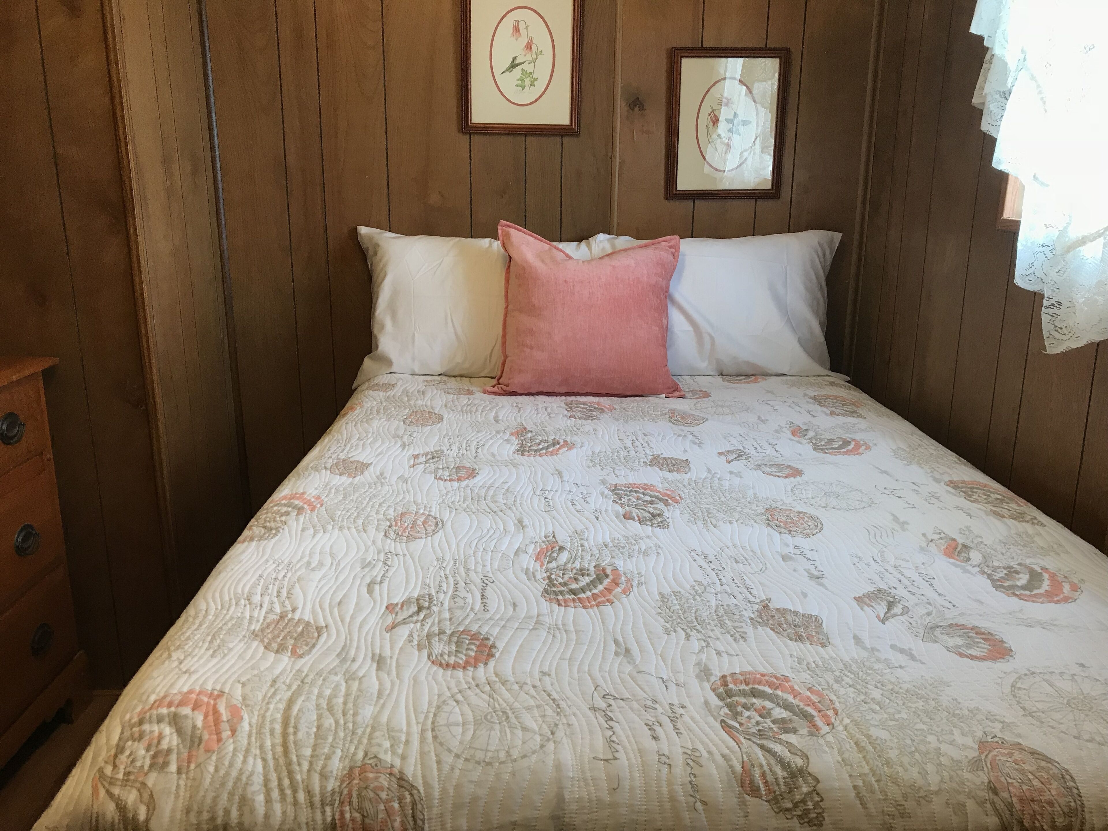 A bedroom with a wooden paneled wall, a bed with a patterned bedspread, two pillows, and two framed pictures on the wall.