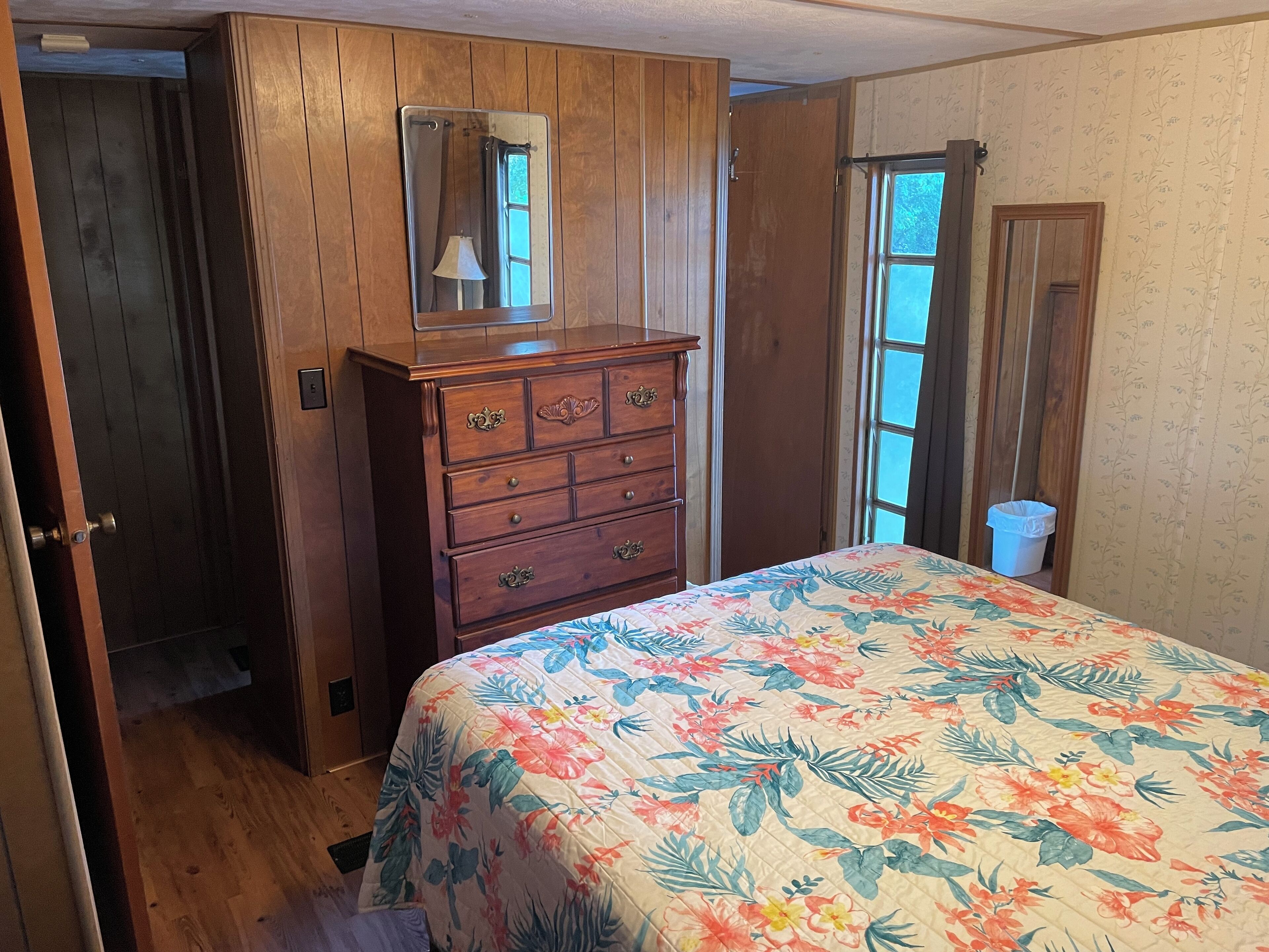 Un dormitorio con cómoda de madera, cama con colcha estampada, ventana con vistas y puerta que da a otra habitación.