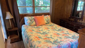 Un dormitorio con edredón estampado con motivos florales, una mesita de noche con lámpara, un aparador y una ventana con vistas a árboles.