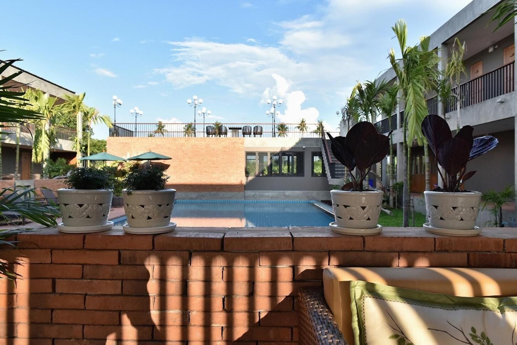 Un espace aménagé au bord de la piscine, agrémenté de plantes en pot, avec un mur en briques et une vue sur un immeuble doté de balcons et d’une piscine.