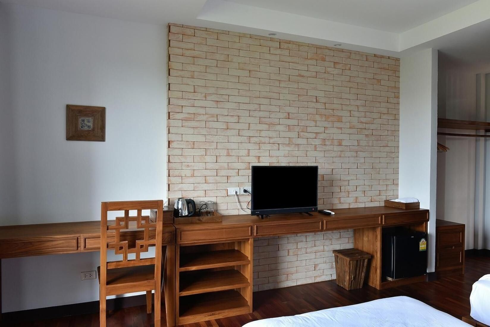 A hotel room with a brick accent wall, a wooden desk with a TV, a chair, a small table with a kettle, and a bed.