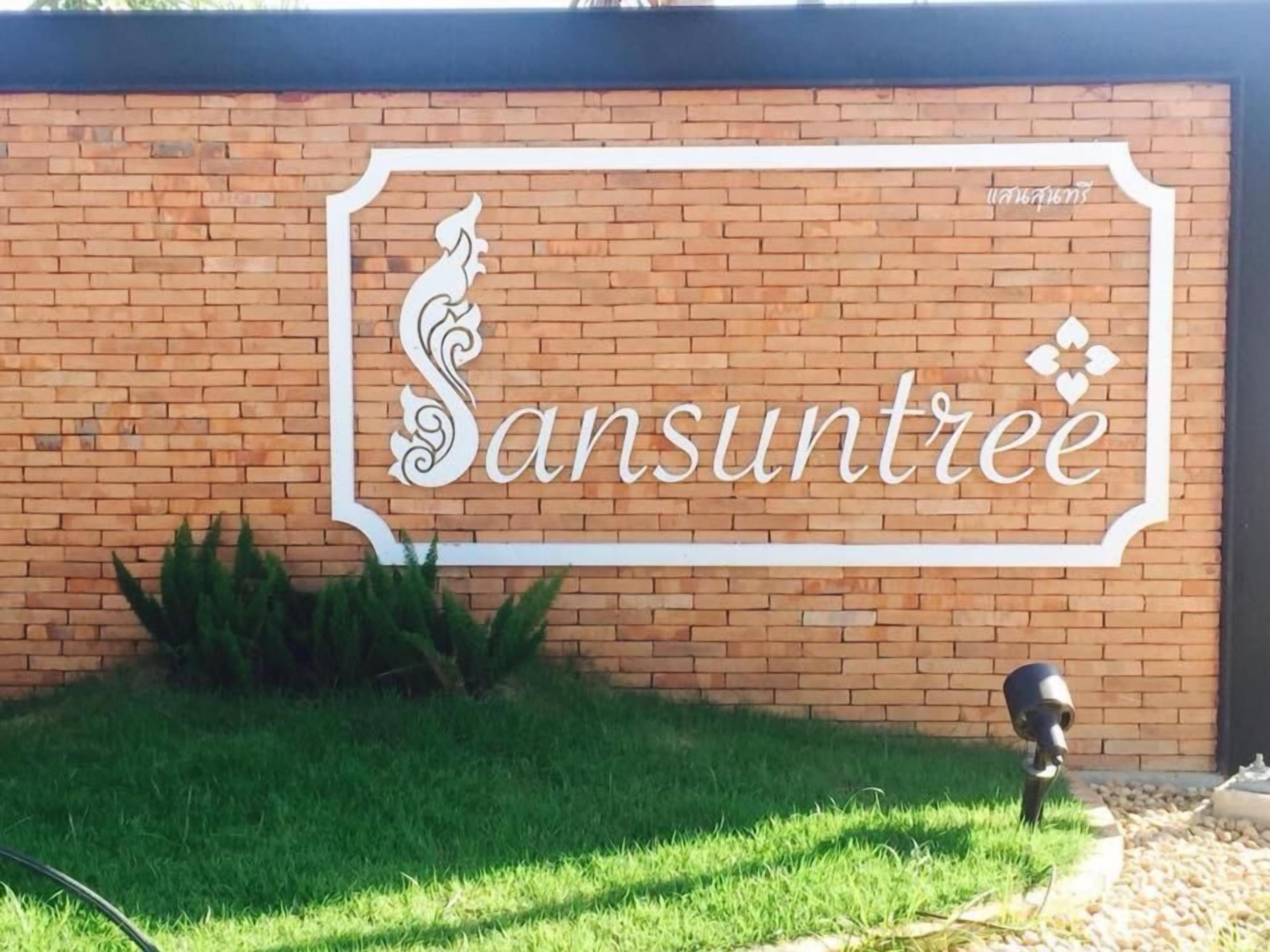 Un muro de ladrillo con un letrero que dice "Sansuntee" en letras estilizadas, rodeado de césped verde y una pequeña zona ajardinada.