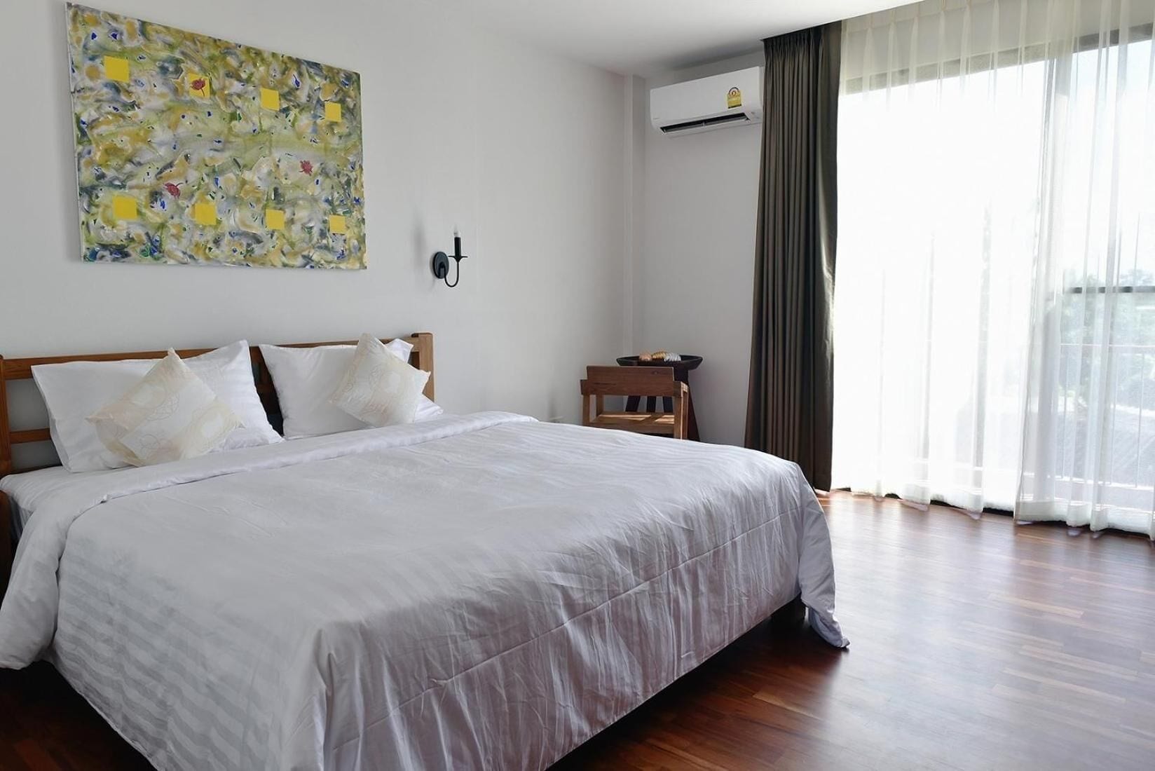 A hotel room with a large bed, a painting on the wall, an air conditioning unit, and a view of the outdoors through sheer curtains.