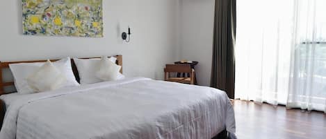 A hotel room with a large bed, a painting on the wall, an air conditioning unit, and a view of the outdoors through sheer curtains.