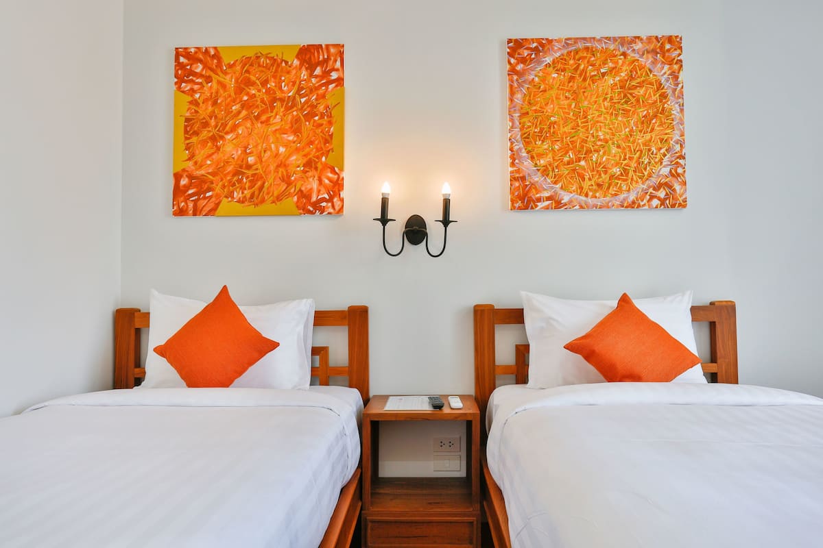 Habitación de hotel con dos camas, cada una con ropa blanca y almohadas naranjas, una mesita de noche con un control remoto y dos cuadros colgados en la pared.