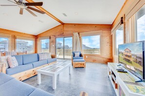 Un vaste espace de vie comprenant un canapé, une table basse et un fauteuil, des murs en bois, de grandes fenêtres et un ventilateur de plafond.