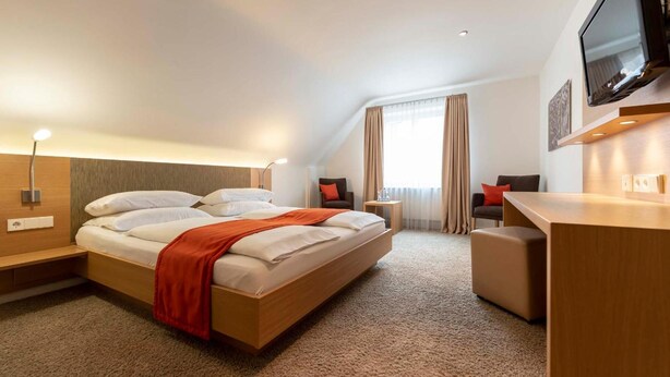 Habitación de hotel con cama, escritorio, silla, televisor y ventana con cortinas.