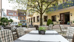 Un área de estar al aire libre con mesas y sillas de metal, una planta en maceta y un edificio con balcón al fondo.