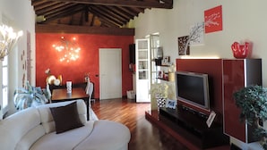 A living room with a wooden ceiling, a white sofa, a TV stand, and a red accent wall. - Luxury apartment located 500 metres from  historic centre and railway station (Varese)