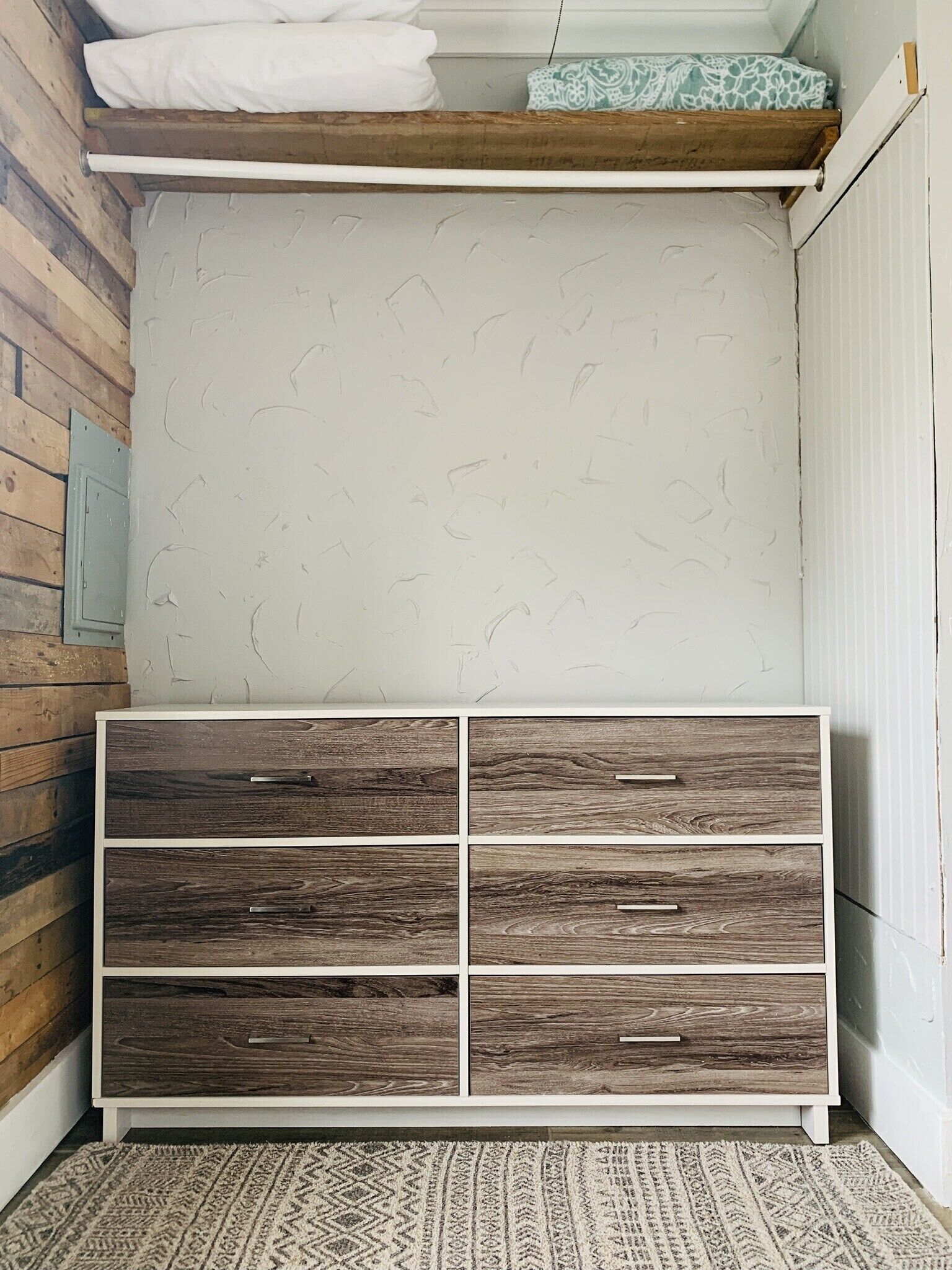 Habitación de esquina con un armario de madera, una alfombra estampada y una pared blanca con textura sutil.