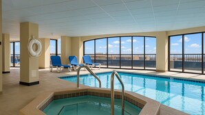 An indoor swimming pool with a hot tub, blue lounge chairs, and large windows offering a view of the sky.