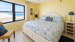 A bedroom with a bed, a window with a view of the beach, a ceiling fan, and a nightstand with a lamp.