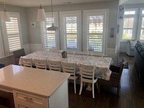 Un comedor con una mesa larga, sillas blancas y un mantel con estampado floral. Hay dos lámparas colgantes sobre la mesa.