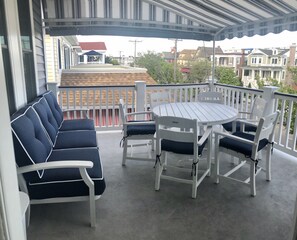 Un balcón techado con una mesa y sillas blancas, un sofá azul y vistas a casas circundantes.