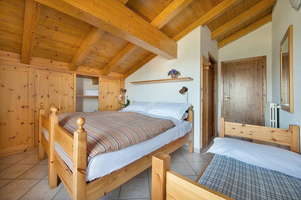A wooden bed with a striped bedspread, a small shelf with a potted plant, and a wooden wardrobe in a room with wooden beams on the ceiling.
