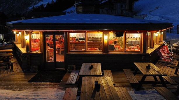 Un edificio de madera con techo cubierto de nieve, ventanas iluminadas y mesas de madera al aire libre.
