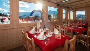 Un comedor con paredes de madera, ventanas amplias y mesas dispuestas para dos, cubiertas con manteles rojos.