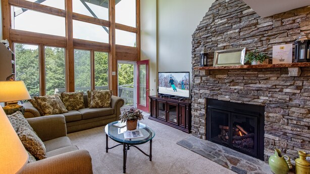 A cozy living room with a stone fireplace, large windows, a comfortable sofa, and a television. - 3B Condo Overlooking Sugar Ski Slopes! (Sugar Mountain)