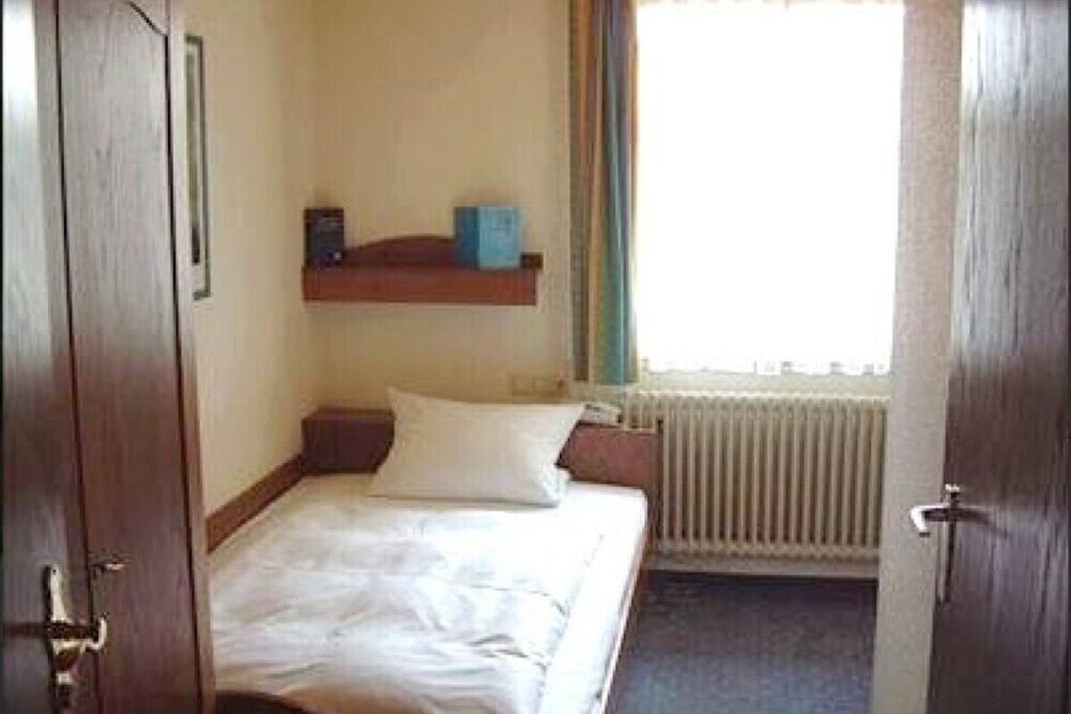 Una habitación pequeña con una cama individual, una puerta de madera, un radiador y una ventana con persianas.