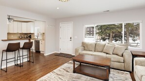 A modern living room with a sofa, coffee table, and bar stools. A kitchen with white cabinets and a countertop is visible in the background. - Beautiful Home in Willits! (Basalt)