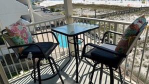 Un balcón con dos sillas y una mesa, con vista a una playa y un paseo marítimo, con gente disfrutando la playa.