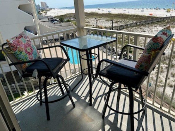 Een balkon met twee stoelen en een tafel, met uitzicht op een strand met een promenade en mensen op het strand.