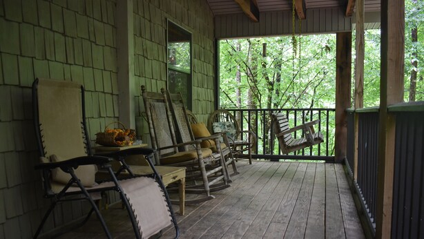 A porch with wooden flooring and a railing, featuring several rocking chairs and a table with a basket.