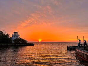A waterfront scene with a lighthouse, calm water reflecting sunset colors, and a dock with chairs. - Walk to beach , bay , shopping & restaurants , Sunset Views at end of our st 1 (Harvey Cedars)