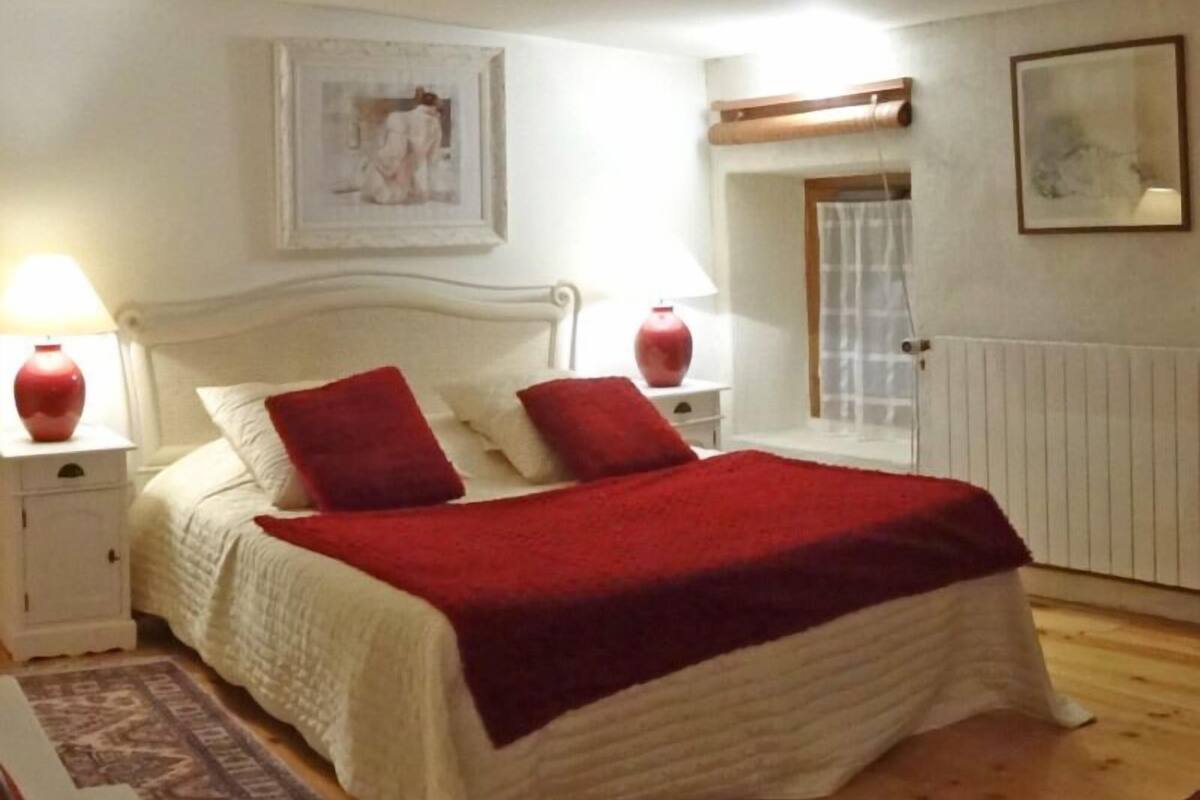 A neatly arranged bedroom with a white bed, red pillows, and a red throw blanket. There are two bedside tables with lamps, a framed picture on the wall, and a window with a view of the outside.