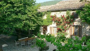 A stone patio with a wooden table and chairs, surrounded by lush greenery and a stone building with a tiled roof. - Le Moulinage (Saint-Thomé)