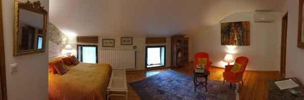Un dormitorio con cama, una silla, una mesa, un ventanal y un cuadro en la pared.