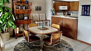 A dining area with a round table, chairs, and a rug. There is a kitchen area with wooden cabinets and a microwave. A potted plant is visible in the corner. - Relaxing Marvelous Main Channel View! (Lake Ozark)
