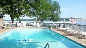 A rectangular swimming pool with a metal ladder in the foreground, surrounded by lounge chairs and a view of a body of water with an American flag. - Relaxing Marvelous Main Channel View! (Lake Ozark)