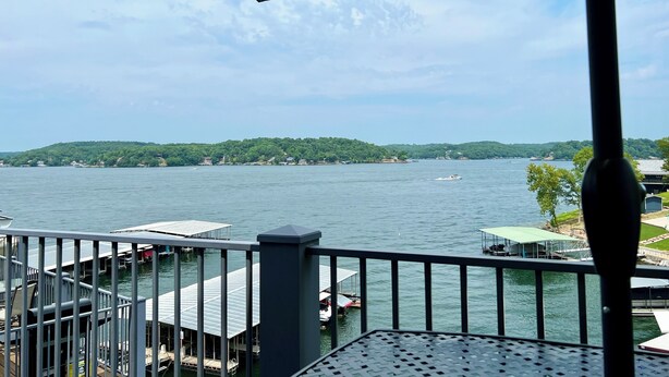 A view from a balcony overlooking a lake with docked boats, a pier, and a distant green hill. - Relaxing Marvelous Main Channel View! (Lake Ozark)
