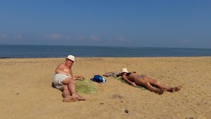 Deux personnes se prélassent au soleil sur une plage de sable, avec un ciel dégagé et une mer calme en arrière-plan.
