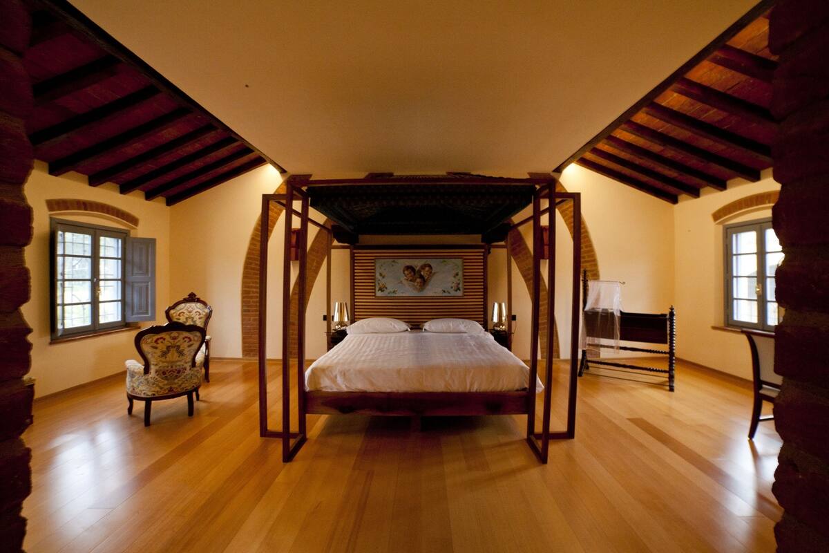 Ein Schlafzimmer mit einem Himmelbett, Holzboden, einem Sessel und einem Bild an der Wand.