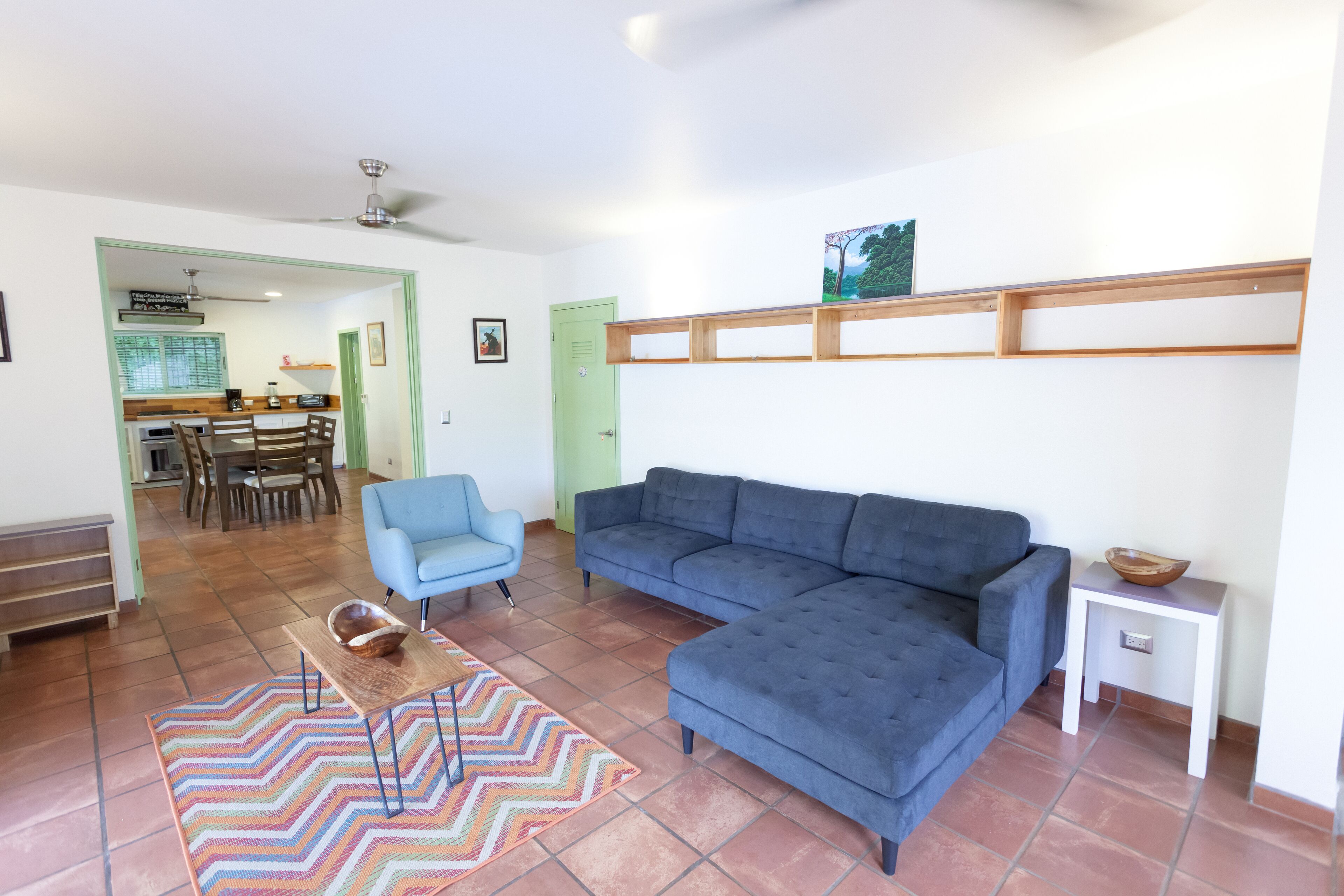 A living room with a blue sofa, a blue armchair, a wooden coffee table, and a colorful rug. There is a kitchen in the background with a dining table and chairs.
