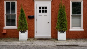 A red textured building with a white door, two windows, and two potted evergreen trees. - 6 Bells: Harbor Springs Lodging for the Discerning Traveler (Harbor Springs)