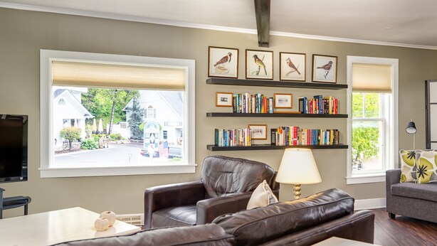 A living room with a brown leather sofa, a white coffee table, a grey armchair, and a bookshelf filled with books. There are two windows with blinds, a lamp on the coffee table, and a framed picture of birds on the wall. - 6 Bells: Harbor Springs Lodging for the Discerning Traveler (Harbor Springs)