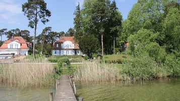 A wooden dock leading to a calm lake with tall grasses, surrounded by lush greenery and two large houses with red roofs.