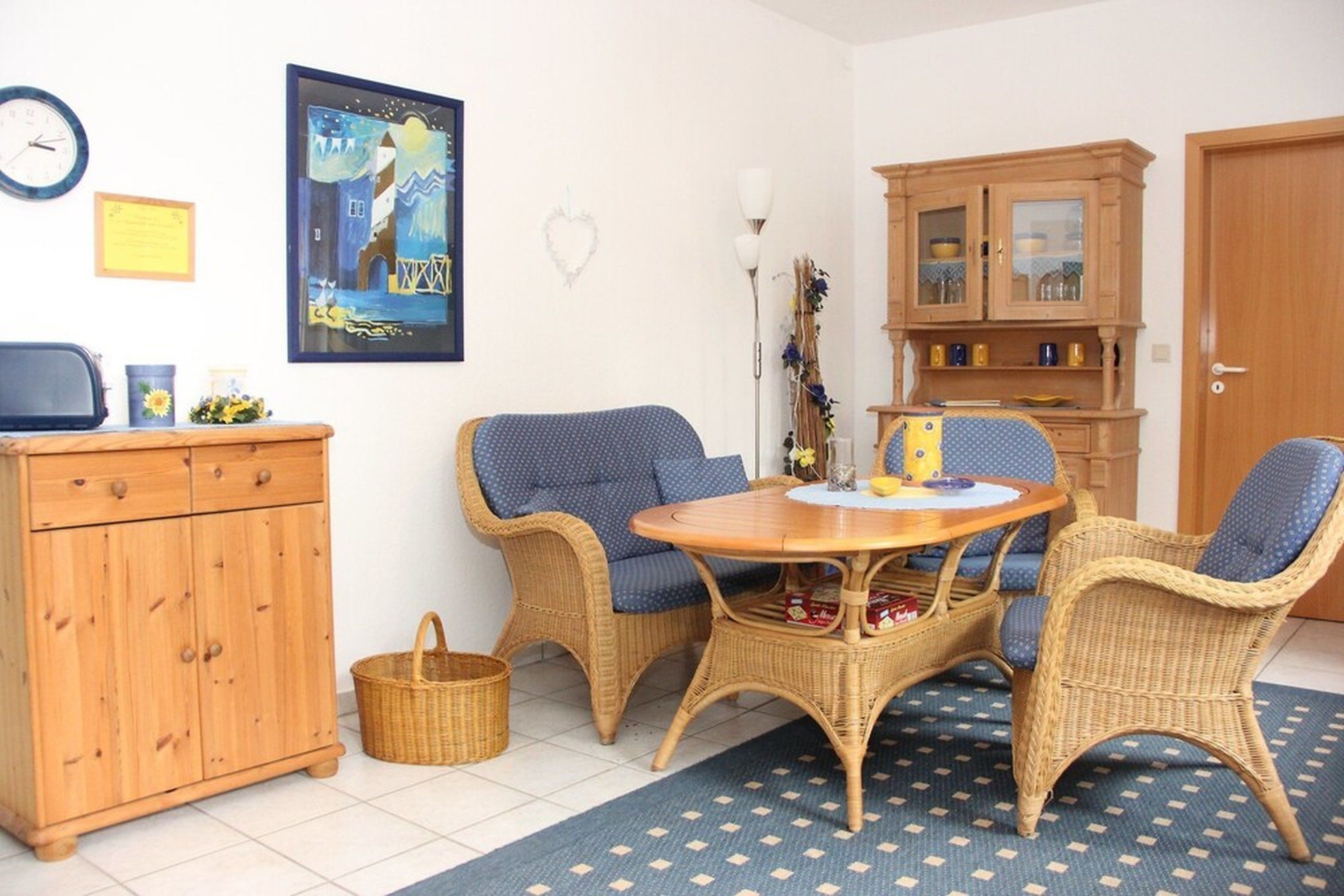 Una habitación con un armario de madera, sillas de mimbre, una mesa redonda y una alfombra azul estampada.
